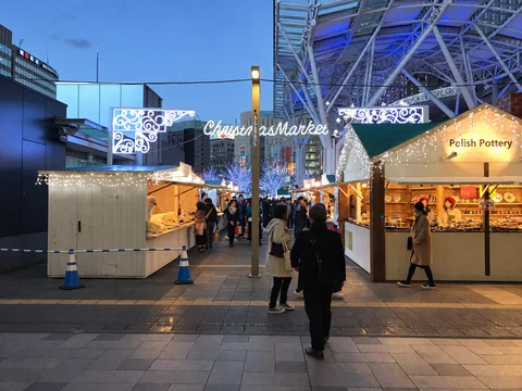 AI-generated representative illustration. Reference photograph: Christmas market stalls and festive lights in front of Hakata Station in Fukuoka. Archival photograph from 2018; the past display is shown.