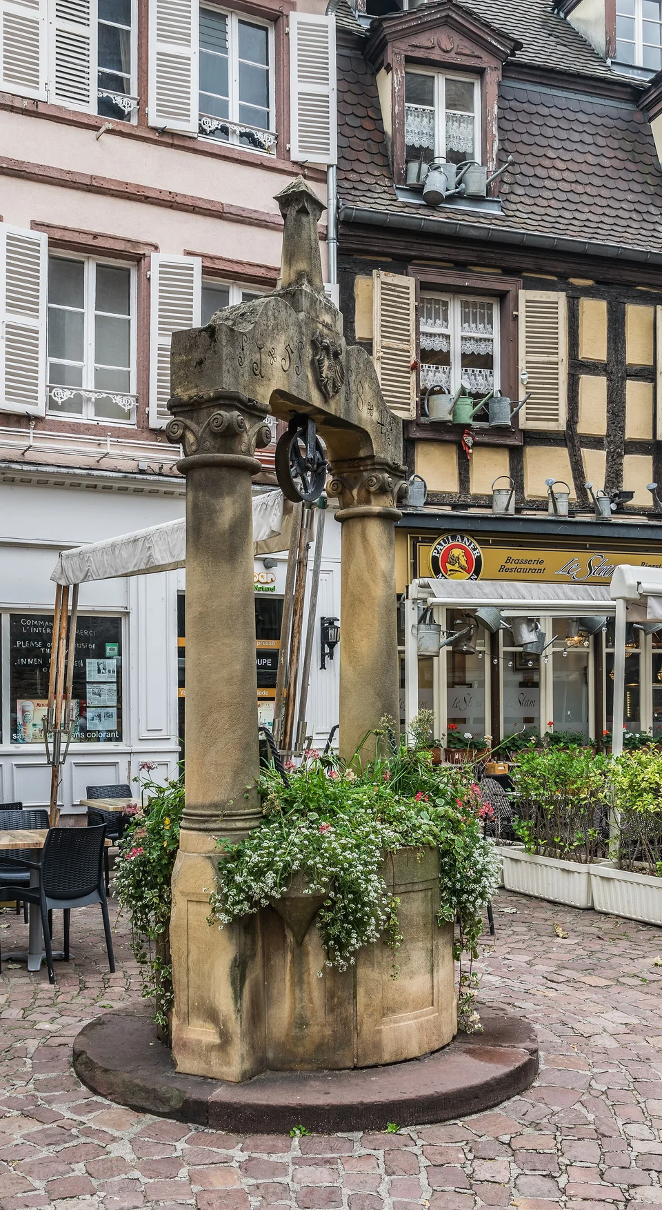 Steinbrunnen mit Blumen und Fachwerkhäusern im Hintergrund am Place des Dominicains in Colmar. Aufnahme des Veranstaltungsorts aus 2018 außerhalb der Weihnachtszeit; sie zeigt nicht den Markt von 2026.