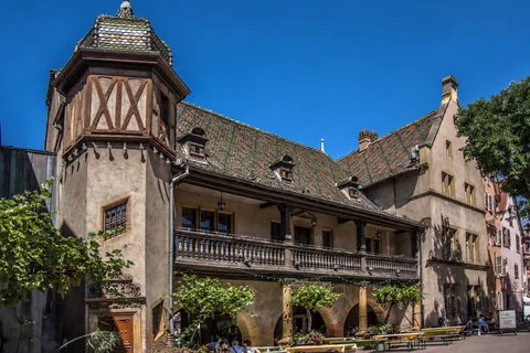 KI-generierte repräsentative Illustration. Fotovorlage: Ziegeldach, Fachwerktürmchen und Balkon des Koïfhus vom Place de l’Ancienne Douane in Colmar aus gesehen. Aufnahme des Veranstaltungsorts aus 2016 außerhalb der Weihnachtszeit; sie zeigt nicht den Markt von 2026.