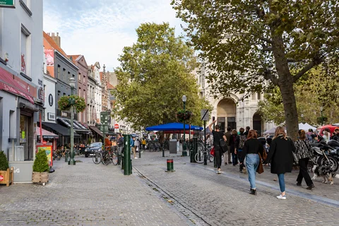 KI-generierte repräsentative Illustration. Fotovorlage: Fußgänger und Bäume auf dem Weg von der Rue du Vieux Marché aux Grains zum Place Sainte-Catherine in Brüssel. Aufnahme des Veranstaltungsorts aus 2019 außerhalb der Weihnachtszeit; sie zeigt nicht den Markt von 2026.