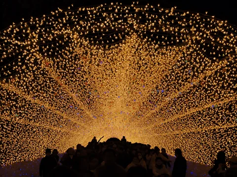 Ilustración representativa generada con IA. Fotografía de referencia: Visitantes caminando por un túnel de luces cálidas en Nabana no Sato. Fotografía de archivo de 2011; muestra una decoración anterior.