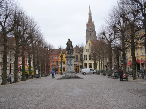 KI-generierte repräsentative Illustration. Fotovorlage: Simon-Stevin-Statue, kahle Bäume und Turm der Liebfrauenkirche am Simon Stevinplein in Brügge. Aufnahme des Veranstaltungsorts aus 2005 außerhalb der Weihnachtszeit; sie zeigt nicht den Markt von 2026.
