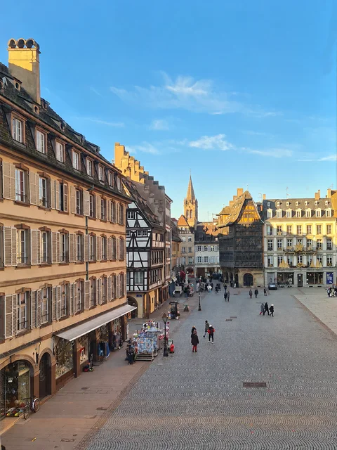 KI-generierte repräsentative Illustration. Fotovorlage: Erhöhter Blick auf das Kopfsteinpflaster des Place de la Cathédrale, Fachwerkfassaden und die Touristeninformation in Straßburg. Archivaufnahme des Ortes aus 2022; sie dokumentiert nicht den Weihnachtsmarkt von 2026.