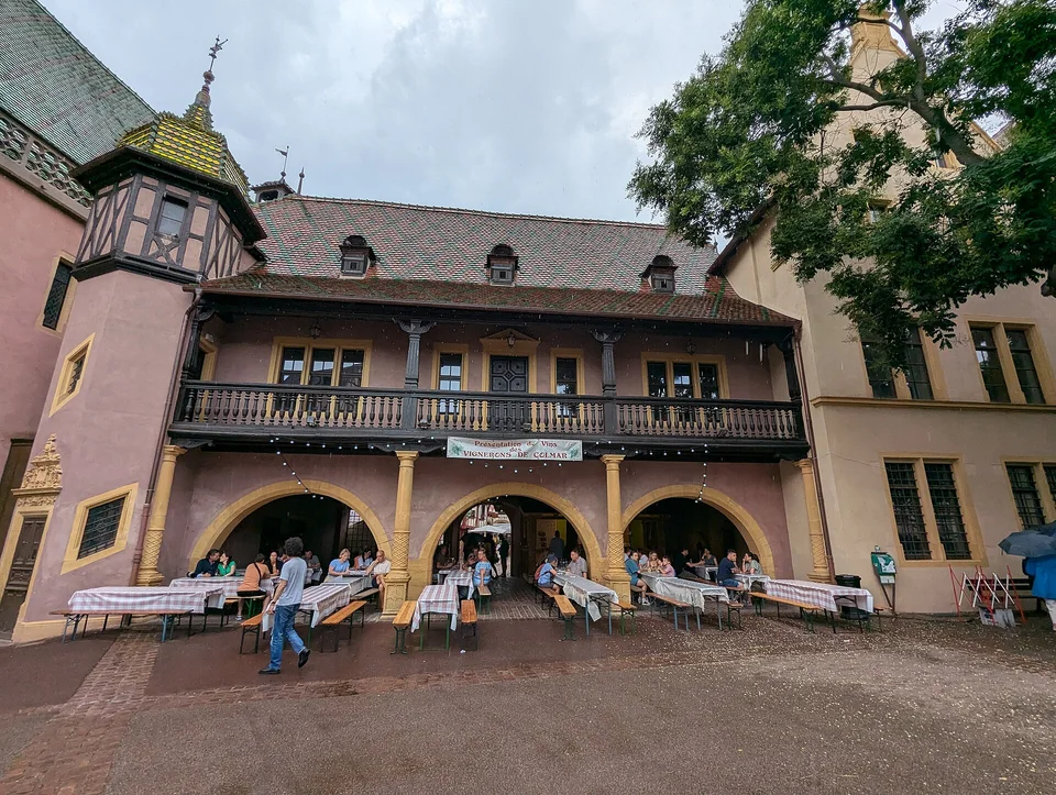 Ostfassade des Koïfhus mit Bögen, Holzbalkon und Außentischen am Place de l’Ancienne Douane in Colmar. Aufnahme des Veranstaltungsorts aus 2024 außerhalb der Weihnachtszeit; sie zeigt nicht den Markt von 2026.