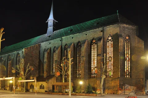 KI-generierte repräsentative Illustration. Fotovorlage: Dominikanerkirche mit beleuchteten Glasfenstern bei Nacht am Place des Dominicains in Colmar. Aufnahme des Veranstaltungsorts aus 2010 außerhalb der Weihnachtszeit; sie zeigt nicht den Markt von 2026.