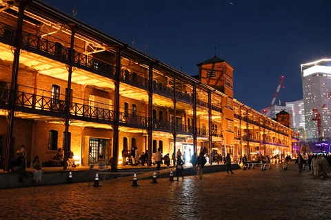 Ilustración representativa generada con IA. Fotografía de referencia: Exterior y balcones iluminados de Yokohama Red Brick Warehouse de noche. Fotografía de archivo de 2021 para situar los almacenes; no muestra puestos del mercado.