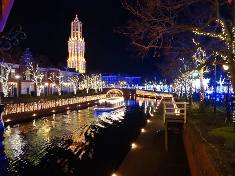 Ilustración representativa generada con IA. Fotografía de referencia: Torre, puente y canal iluminados de Huis Ten Bosch, Sasebo, de noche. Fotografía de archivo de 2012; muestra una decoración anterior.